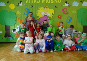 Dzieci z grupy III w wiosennych przebraniach z nauczycielką, na tle dekoracji.